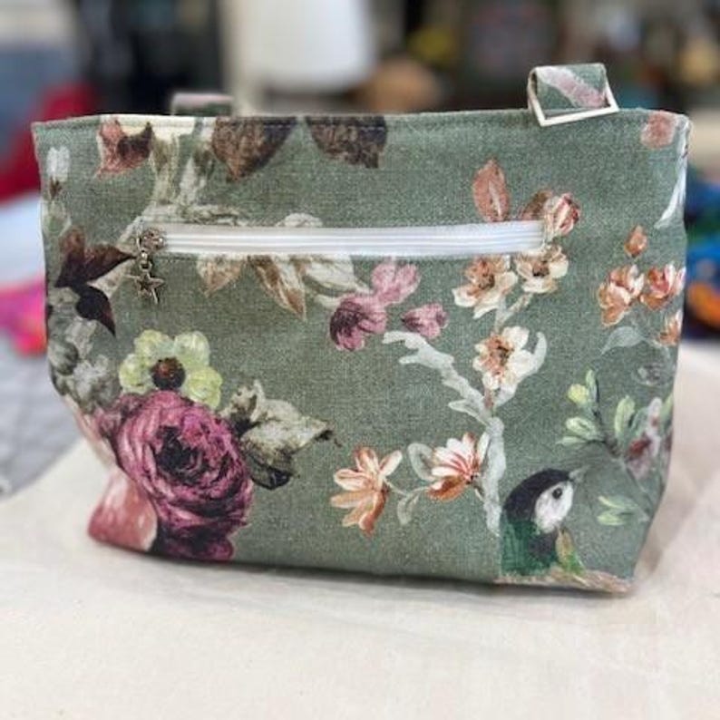 A sage green fabric bag with a floral pattern. The design features pink, white, and brown flowers, a white zip, and a small star-shaped charm. The bag has a small handle and a floral design that includes a bird. The bag was made using the Phillippa Crossbody sewing pattern.
