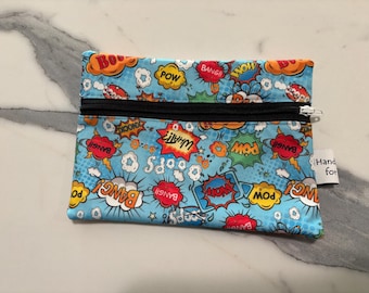 May include: A rectangular fabric zipper pouch featuring a light blue background with a colorful comic book pattern. The design includes various speech bubbles with text such as POW, BANG, and WOW in red, yellow, and orange. A black zipper runs across the top.