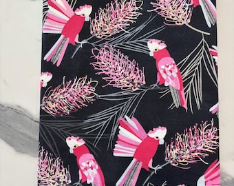 May include: A rectangular fabric book cover featuring a black background with a repeating pattern of pink and white cockatoos perched on branches with pink bottlebrush flowers and thin grey leaves. A small yellow ribbon bookmark extends from the bottom.