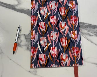 May include: A rectangular notebook with a dark blue cover featuring a repeating pattern of pink, red, and white protea flowers. A silver pen with an orange grip is positioned to the left of the notebook, and a solid pink ribbon bookmark hangs from the bottom.