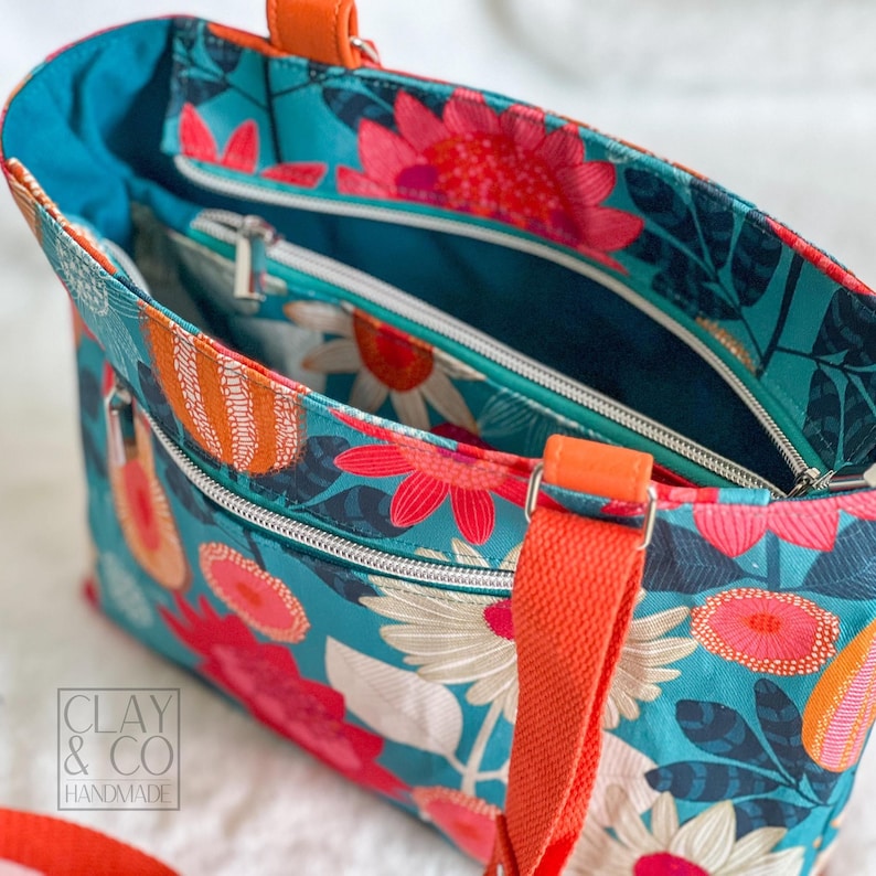 A floral tote bag with a teal background and large flowers in shades of pink, orange, and white. The bag has orange straps and handles, and silver zippers. The interior is teal. The bag is approximately 30 cm wide and 25 cm tall. The bag was made using the Phillippa Crossbody sewing pattern.