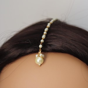 May include: A gold-toned forehead ornament, or maang tikka, featuring a delicate chain of small pearls. A larger, pear-shaped pearl pendant hangs at the forehead. The ornament is set against dark brown hair and a neutral background.