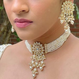 Puede incluir: Un collar gargantilla de perlas blancas con un colgante dorado con un diseño floral y detalles de perlas. El colgante es una pieza grande y adornada con una piedra central rodeada de piedras más pequeñas.