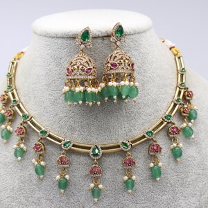Puede incluir: Juego de collar y pendientes dorados con detalles de piedras preciosas verdes y rojas. El collar y los pendientes presentan un diseño detallado con cuentas verdes colgantes y pequeñas perlas blancas. Los pendientes tienen forma de campana.