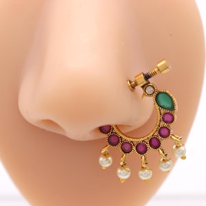 May include: A gold-toned nose ring with a decorative design. It features a green and red gemstone, along with small white pearls. The nose ring is attached to a nose with a screw-like mechanism.