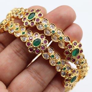 May include: Two gold bangles with floral designs. Each bangle features emerald green, ruby red, and clear gemstones. The bangles are held in a hand against a white background. The jewelry is ornate and detailed.