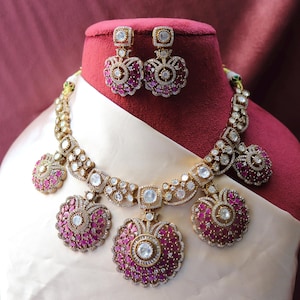 May include: A gold necklace with a pink and white stone design. The necklace has a pendant with three large, round, pink stones surrounded by smaller white stones. The necklace is displayed on a red velvet mannequin bust.
