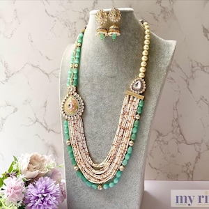 Può includere: Una collana multifilo con un ciondolo a forma di lacrima, composta da perle bianche e verdi. La collana è abbinata a orecchini.