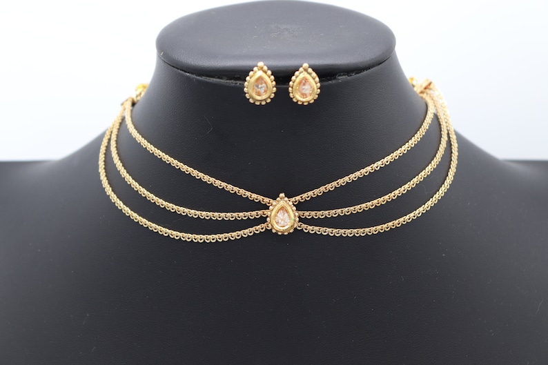 Pu&ograve; includere: Un girocollo dorato con tre catene delicate e un ciondolo a forma di lacrima con una pietra trasparente. Orecchini a goccia abbinati con pietre trasparenti.