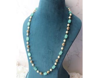 May include: A long necklace with alternating light blue, white, and gold-colored beads. The necklace is displayed on a teal mannequin. The beads are rectangular, round, and spherical, creating a layered effect. The necklace is a fashion accessory.
