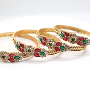 May include: Four gold-colored bangles with floral designs. Each bangle features a repeating pattern of red, green, and clear gemstones. The bangles are set against a white background, highlighting the jewelry's intricate details.
