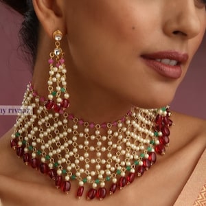 Op de afbeelding: Close-up van een gouden ketting en bijpassende oorbellen. De sieraden hebben witte parels, rode en groene kralen en een open ontwerp. De tekst "my rivaaz" is zichtbaar.