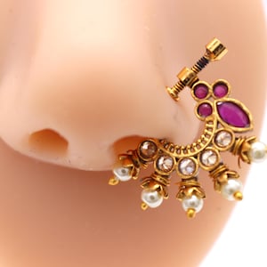 May include: A gold-toned nose ring with a decorative design. The jewelry features clear and purple gemstones, along with small white pearls. The nose ring is designed to be worn on the side of the nose.