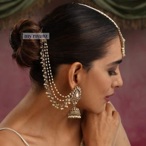 May include: A woman wearing a traditional Indian headpiece and earrings with pearl and gold accents. The earrings are jhumka style with a dangling pearl and a chain attached to the ear. The headpiece is a maang tikka with a pearl chain and a pendant.
