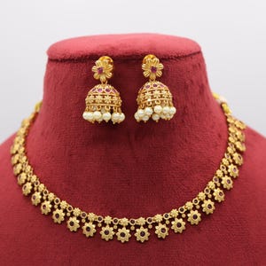May include: A gold-coloured jewellery set comprising a necklace and matching earrings. The necklace and earrings feature a floral design with red accents. The earrings are bell-shaped and adorned with small white pearls. The set is displayed on a red velvet bust.