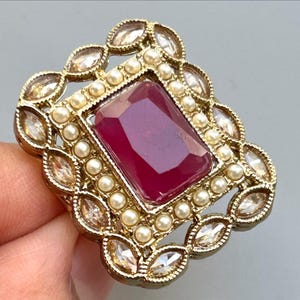May include: A gold-toned ring featuring a large, rectangular, deep red gemstone at its center. The gemstone is surrounded by small, round, white pearls and clear, oval-shaped crystals. The ring has an ornate, vintage-inspired design.