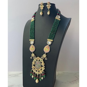 May include: A long green beaded necklace with a large, ornate pendant featuring a pink and green design. The pendant is surrounded by smaller green beads and gold accents. The necklace is paired with matching earrings with pink and green stones.