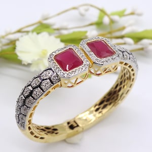 May include: A gold and silver-toned hinged bracelet with two rectangular, ruby-colored gemstones. The gemstones are surrounded by small, clear stones. The bracelet has a textured design on the sides and a patterned interior.