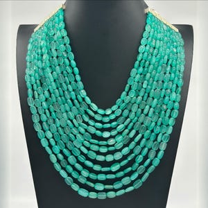 May include: A multi-strand necklace featuring numerous oval-shaped, light-green beads. The necklace is displayed on a black mannequin, with a gold-colored clasp at the top. The beads are uniform in size and color, creating a luxurious look.