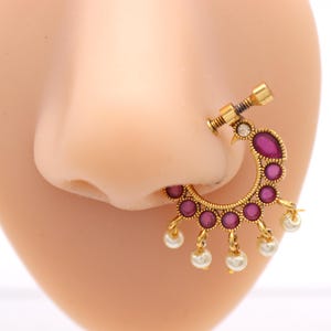 May include: A gold-colored nose ring with a crescent-shaped design. The ring features a series of small, round, magenta-colored stones and dangling white pearls. The nose ring is attached to a nose.
