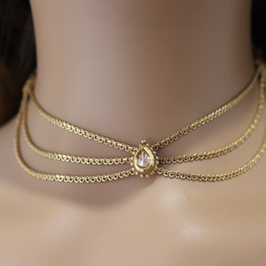 Pu&ograve; includere: Un girocollo dorato con tre catene delicate e un ciondolo di cristallo a forma di lacrima.