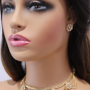 May include: A gold choker necklace with a layered chain design and floral accents. The necklace is adorned with clear stones and has a delicate, feminine look. Matching earrings with a similar floral design are also pictured.