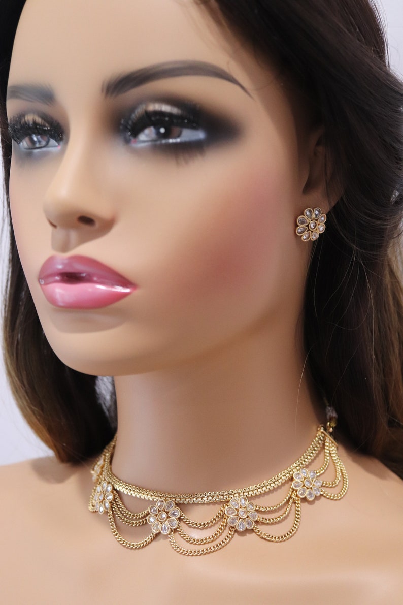 May include: A gold choker necklace with a layered chain design and floral accents. The necklace is adorned with clear stones and has a delicate, feminine look. Matching earrings with a similar floral design are also pictured.