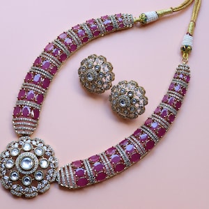 Può includere: Un set di collana e orecchini con un pendente floreale e orecchini abbinati. La collana e gli orecchini sono adornati con pietre color rubino e cristalli trasparenti, incastonati in un metallo dorato. I gioielli sono esposti su uno sfondo rosa chiaro.