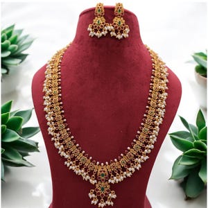 Puede incluir: Juego de collar y pendientes dorados, exhibido sobre un busto rojo. La joyería presenta intrincados diseños florales, pequeñas perlas blancas y detalles en rojo y verde. El collar tiene un colgante con un diseño similar.