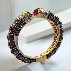 May include: A gold bangle bracelet with a black enamel design and red gemstones. The bracelet is hinged and has a decorative clasp.