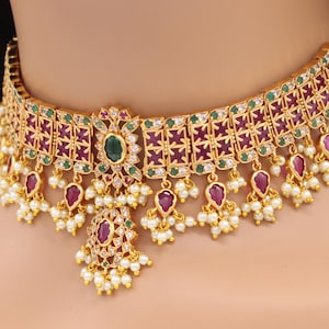 Puede incluir: Un collar gargantilla de oro con un diseño de piedras rojas y verdes y perlas blancas. El collar tiene un diseño floral central con una piedra verde grande.