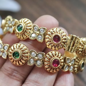 May include: A pair of gold-toned hinged bangles featuring alternating circular motifs with embedded red and green stones, interspersed with four-petaled floral shapes set with clear white crystals, showcasing a textured metallic finish throughout.