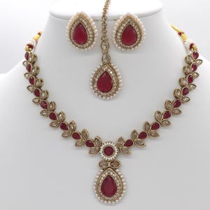 May include: A three-piece jewelry set featuring a necklace, earrings, and a headpiece. The set has a gold-tone metal base with red teardrop-shaped stones and small white pearls. The necklace and headpiece have a floral design.