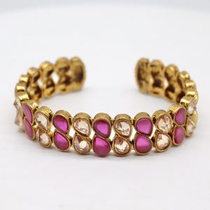 May include: Gold-toned cuff bracelet with alternating pink and clear teardrop-shaped gemstones. The open-ended bracelet features a textured design on the metal band, creating a vintage or antique aesthetic. The gemstones are set closely together.