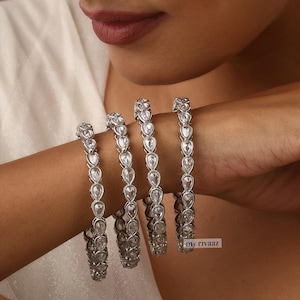 May include: Set of four silver bangles with clear teardrop-shaped stones. The bangles are stacked on a person's wrist.
