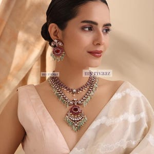 May include: A two-piece jewelry set featuring a necklace and matching earrings. The necklace has a layered design with a large pendant. The earrings and pendant have red and green accents. The text "my rivaaz" is visible.