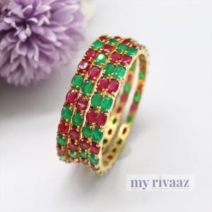 Puede incluir: Tres brazaletes de oro con piedras preciosas rojas y verdes alternas. Los brazaletes están apilados uno encima del otro.