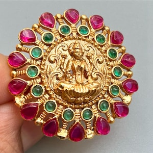 May include: A gold-colored ring featuring a central figure surrounded by alternating teardrop-shaped red and round green gemstones. The ring has an ornate, detailed design.