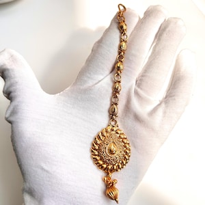 May include: A gold-toned maang tikka with a round, ornate pendant and a dangling teardrop-shaped bead. The tikka is made of a chain with small, round beads.