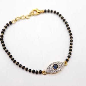 May include: A gold-toned bracelet featuring a central eye-shaped charm with a blue center stone and surrounding clear crystals. The bracelet's chain is composed of small, black beads. The clasp and connecting elements are gold-colored.