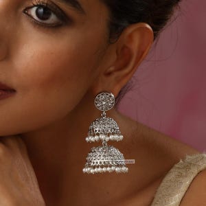 May include: Silver dangle earrings with a layered design. The top layer is a round, filigree design with a pearl in the center. The bottom layer is a bell-shaped design with a row of pearls around the bottom.