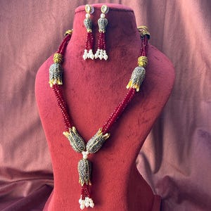 May include: A red and gold necklace with a pendant shaped like a flower. The necklace is made of beads and has a tassel at the bottom. The necklace is displayed on a red velvet stand. Matching earrings are also included.