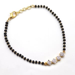 May include: A gold-toned bracelet featuring a chain of small black beads. The bracelet has a gold clasp and is accented with three gold-framed, flower-shaped charms, each set with small, clear crystals. The bracelet is displayed on a white background.