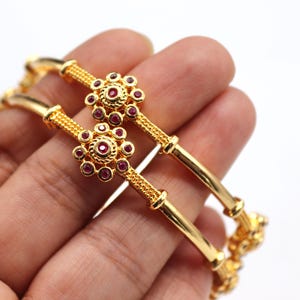 Op de afbeelding: Twee gouden armbanden met bloemaccenten en kleine rode edelstenen. De armbanden worden in een hand gehouden tegen een witte achtergrond. De bloemaccenten hebben een grotere rode edelsteen in het midden.