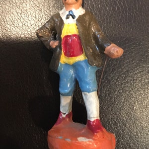 May include: A hand-painted ceramic figurine of a man wearing a brown jacket, yellow vest, red sash, blue pants, and white socks. He is holding a stick in his right hand and has a black top hat on his head.
