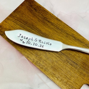 May include: A silver butter knife with the engraved text "Joseph & Nicole 10.10.26" and two small heart symbols. The knife rests on a wooden cutting board with a light pink background.