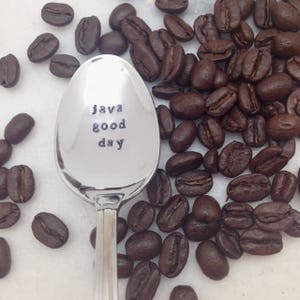 May include: A silver spoon with the words "java good day" engraved on the front. The spoon is surrounded by coffee beans.