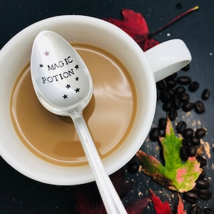 May include: A silver spoon with the words "Magic Potion" and stars engraved on the bowl. The spoon is resting in a white mug filled with coffee. Coffee beans and fall leaves are scattered around the mug.