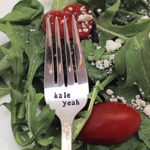 May include: A silver fork with the words "kale yeah" engraved on the handle. The fork is resting on a salad of arugula, tomatoes, and crumbled cheese.