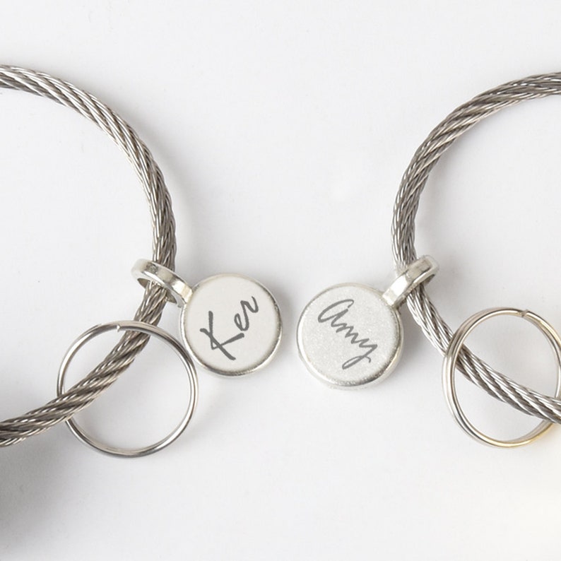Op de afbeelding: Twee zilveren sleutelhangers met gegraveerde namen, "Amy" en "Ker", op ronde labels. De sleutelhangers zijn gemaakt van gevlochten metalen kabel.